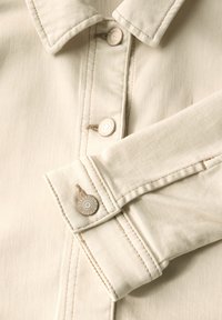 Camicia beige chiaro con colletto classico, dotata di due chiusure a bottone e un tessuto testurizzato con cuciture a contrasto e accessori metallici firmati.