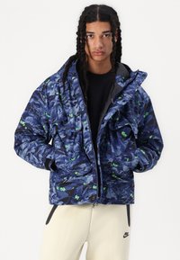 Puffer jacket in navy with green leaf pattern, featuring zip and snap closures, multiple pockets, and textured finish. Worn over a black top.