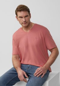 T-shirt z dekoltem w serek w jasnym koralowym kolorze, wykonany z miękkiego materiału. Posiada krótkie rękawy i subtelne logo na piersi, noszony z niebieskimi dżinsami.