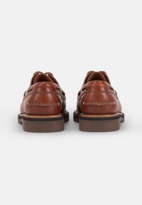 Chaussures en cuir marron avec une forme arrondie, présentant des découpes cousues et une semelle noire contrastante. La vue arrière met en évidence le laçage et la texture.