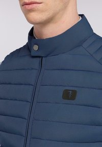 Námořnická modrá prošívaná bunda se zipem, stojáčkovým límcem a černou nášivkou. Disponuje horizontálními prošívanými pruhy a kovovými patentky.