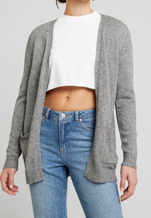 Femme portant un cardigan gris ouvert sur un t-shirt court blanc et un jean bleu taille haute sur un fond uni.