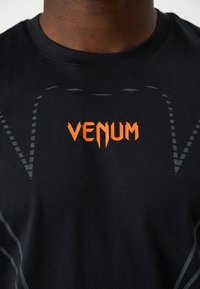 Camisa atlética de tejido negro con un logo "VENUM" naranja y sutiles patrones en gris, que presenta un diseño ajustado y una textura suave.