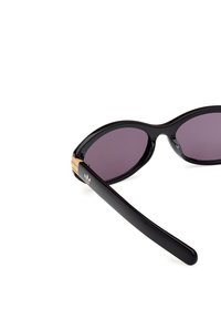Zwarte zonnebril met een glanzende afwerking, paarse lenzen en een gouden accent op de veren met een logo. Slank, ovaal frame.