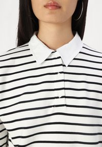 Polo shirt à col haut avec des rayures horizontales noires sur un fond blanc. Il présente une patte de boutonnage et une texture de tissu lisse.