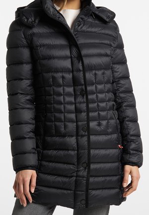 Femme portant une doudoune noire matelassée mi-longue avec capuche et boutons-pression sur un top en maille blanche.