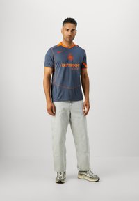 Maillot de sport Nike gris avec des accents orange et un logo, présentant des manches courtes et une coupe décontractée, associé à un pantalon en denim clair et des baskets.
