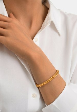 Bracelet en or tressé avec une finition lisse et polie, porté au poignet, complétant une chemise blanche à col.