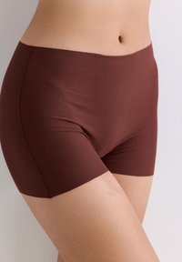 Close-up van het middel van een persoon die naadloze bruine boyshort onderbroek draagt, met zicht op de taille, heup en bovenbeen tegen een neutrale achtergrond.