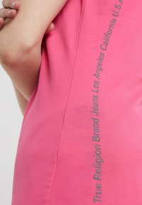 Camiseta ajustada de color rosa hecha de algodón suave. Presenta texto vertical en gris en el lado: "True Religion Brand Jeans Los Ángeles California EE.UU."