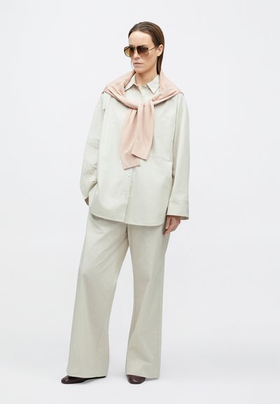 Világos bézs, hosszított ing és hozzá illő bő szárú nadrág, vállra terített halvány rózsaszín pulóverrel és barna napszemüveggel.