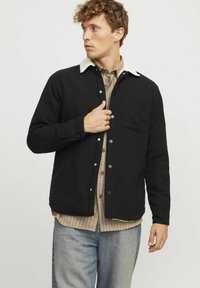 Zwarte button-up jas met een beige schapenwollen kraag, voorzien van een voorkantzak en een relaxte pasvorm, gedragen over een geruite overhemd.