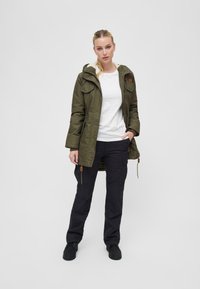 Olive green parka jacket with a hood, front button closure, and two chest pockets, paired with black cargo pants and black shoes.