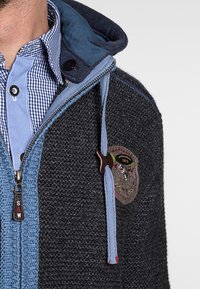 Grijze en blauwe wollen jas met een rits aan de voorkant, capuchon, textuur gebreid en een decoratieve patch met geborduurde details.