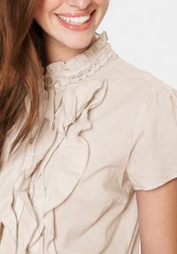 Beige Bluse mit Rüschendesign, hohem Ausschnitt und Knopfakzenten. Aus leichtem Stoff gefertigt, mit subtiler Textur und sanfter Drapierung.
