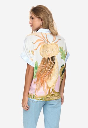 Femme aux longs cheveux blonds portant un t-shirt à manches courtes avec un grand motif détaillé de chouette et de feuillage dans le dos, assorti à un jean bleu clair.