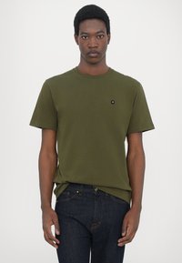 Tricou din bumbac de culoare verde măslin, cu mâneci scurte, guler rotund și un mic logo negru pe piept; asortat cu blugi negri din denim.
