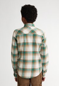 Kostkovaná košile s dlouhým rukávem v krémové, hnědé a tyrkysové barvě. Má límec na knoflíky a zaoblený lem. Vyrobena z měkké tkaniny s kostkovaným vzorem.