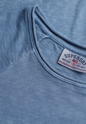 Lyseblå bomulds t-shirt med rund halsudskæring, der har en glat tekstur og en kontrasterende stofdetalje på skulderen. Label synlig.