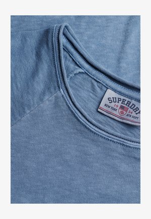 Hellblaues Baumwoll-T-Shirt mit rundem Ausschnitt, glatter Textur und kontrastierendem Stoffdetail an der Schulter. Etikett sichtbar.