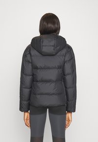 Giacca a vento nera con cappuccio, caratterizzata da un design trapuntato e polsini elasticizzati. Indossata sopra leggings scuri aderenti, creando una silhouette slanciata.