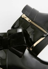 Schwarze Lackankelstiefel mit einem Samt-Schleifendetail, goldenen Reißverschluss und schwarzer Gummisohle. Glatte Oberfläche mit glänzendem Finish.
