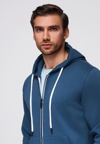 Granatowy bluza z zamkiem błyskawicznym i białymi sznurkami, z przednią kieszenią i miękką teksturą. Nosi się ją na jasnoniebieskiej koszulce, o dopasowanym kroju.