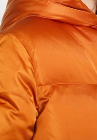 Puffjacka i en glänsande bränd orange färg, med en slät textur, vadderad design och hög krage för extra värme.