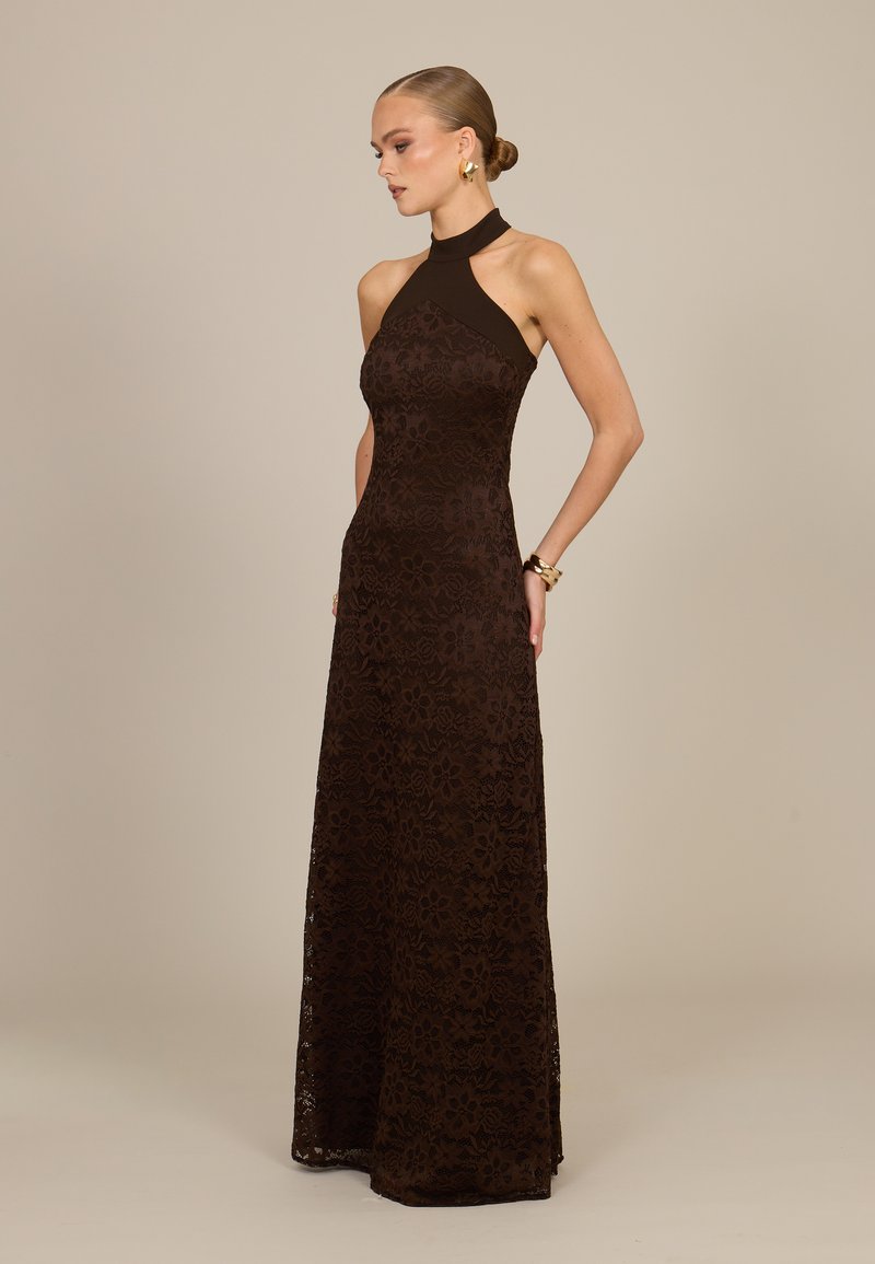 Femme avec les cheveux en chignon portant une robe sans manches à col halter en dentelle florale marron foncé, debout les mains dans le dos devant un fond uni.