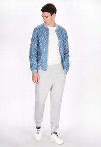 Lichtblauwe bomberjack met een bloemenpatroon, gedragen over een witte T-shirt en grijze joggingbroek. Beschikt over geribde manchetten en een ritssluiting.