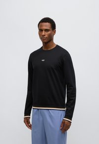 Juodas ilgomis rankovėmis megztinis su balta logotipu ant krūtinės, kontrastingos dryžuotos rankogaliai ir apačia, derinamas su šviesiai mėlynomis kelnėmis. Lygios audinio tekstūros.