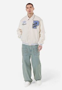 Hvit varsity-jakke med blå broderte bokstaver og detaljer, sammen med lys denim baggy jeans med dekorativ søm. Hvite sneakers.