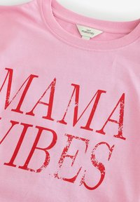 Roze katoenen t-shirt met grote rode tekst "MAMA VIBES." De stof heeft een gladde textuur en een ronde hals.