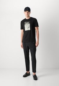 Tricou negru cu un print grafic în centrul său, asociat cu pantaloni negri, tapered și mocasini negri. Material mat cu o textură netedă.