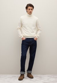 Cremefarbener Rollkragenpullover, dunkelblaue Jeans mit geradem Bein, braune Schnürstiefel. Glatte Textur, klassischer Schnitt, leicht aufgerollte Bündchen, minimalistisches Design.