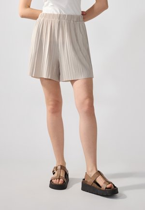 Beżowe spodenki w plisy z elastycznym pasem, zestawione z brązowymi sandałami na paski z masywną czarną podeszwą.
