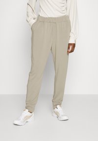 Calças jogger bege com um cós elástico, bolsos laterais e pernas ajustadas, combinadas com sapatilhas brancas de desporto com detalhes em dourado.