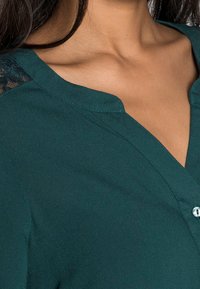 Blouse bleu sarcelle avec un décolleté en V profond, ornée de détails en dentelle sur les épaules et une fermeture à boutons. Texture douce avec un léger brillant.