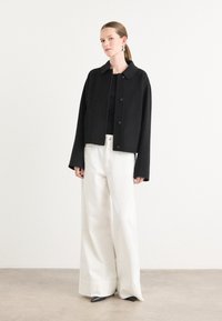 Zwarte cropped jacket met een omgeslagen kraag en knoopsluiting, gecombineerd met wijde witte broeken en puntige zwarte schoenen.