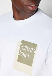 Sudadera de algodón blanca con un gráfico en capas en oliva y beige y el texto "Calvin Klein" en una fuente moderna. Textura suave.