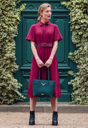 Bordeauxfarbenes Hemdkleid mit kurzen Ärmeln, durchgehender Knopfleiste und geflochtenem Gürtel. Kombiniert mit einer dunkelgrünen gesteppte Tragetasche und schwarzen Stiefeletten.