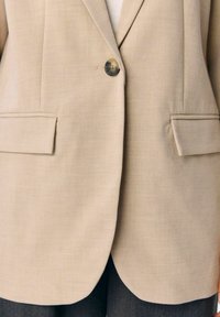 Beige kavaj med en enkel knappstängning, två framfickor och slät tyg med en subtil textur, kompletterad med rundade fållar.