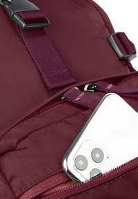 Smartphone bianco parzialmente inserito nella tasca anteriore con cerniera di uno zaino bordeaux con spallacci regolabili e fibbie.