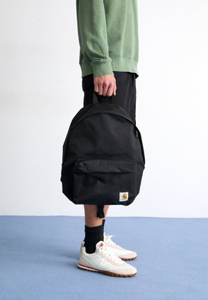 Zaino in tessuto nero con manico superiore, tasca frontale con zip e patch con logo. Indossato da una persona in abbigliamento casual e sneakers.