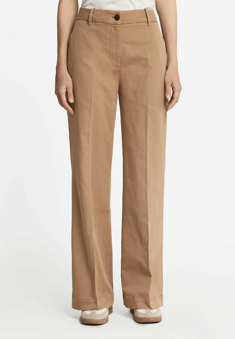 Beżowe spodnie z szerokimi nogawkami wykonane z bawełny o gładkiej fakturze. Mają z przodu zapięcie na guzik i zamek błyskawiczny; noszone z jasnymi sneakersami.