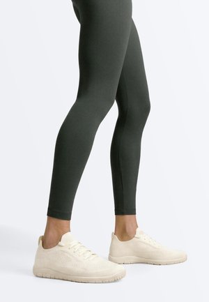Benen met donkergroene leggings en witte gebreide sneakers met veters tegen een effen lichte achtergrond.