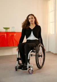 Wheelchair with metallic frame and purple accents. User wears black top and pants, seated indoors with light background and furniture.