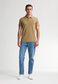 Uomo che indossa una polo beige, jeans azzurri chiari e sneaker bianche, in piedi contro uno sfondo bianco uniforme.