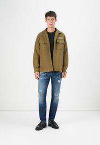 Olivengrøn skjortejakke med to brystlommer, sort T-shirt under, parret med slidte blå jeans og sorte sko.