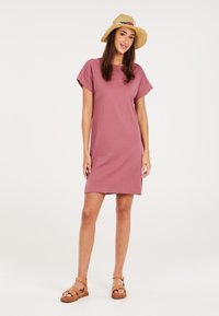 Lässiges kurzärmeliges Kleid in hellem Mauve, aus weichem Stoff gefertigt, mit weiter Passform und seitlichen Schlitzen, kombiniert mit einem Strohhut und Sandalen.